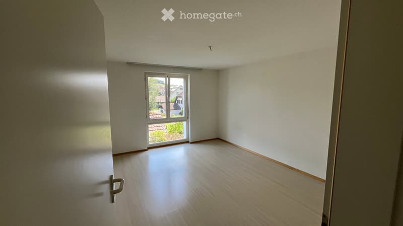 Empty room with light-colored wooden flooring, white walls, and a large window with a view of the outside.