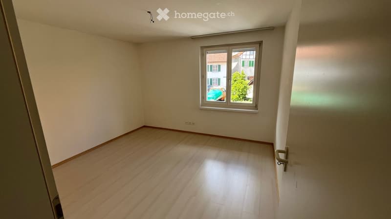 Empty room with wooden flooring, white walls, and a single window showing a view of a neighboring building and greenery.