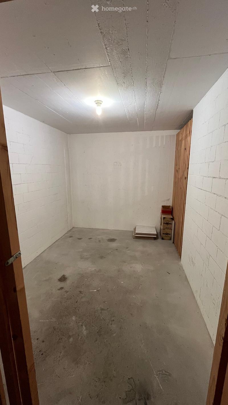 Empty room with white brick walls, concrete floor, and a single light fixture on the ceiling. There are a few boxes and a stack of papers in the corner.