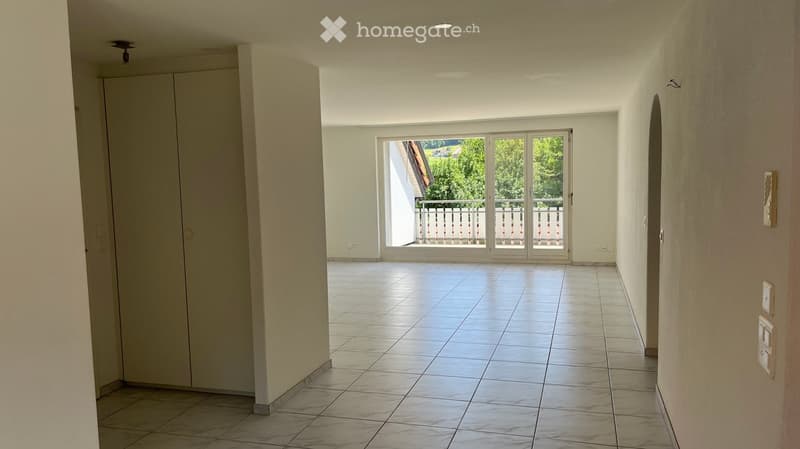 Empty hallway with tiled floor, white walls, and a large sliding glass door leading to a balcony with a view of greenery.