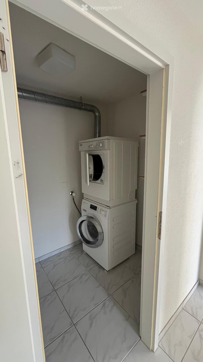 Small utility room with a washing machine and dryer stacked vertically, tiled floor, white walls, and a door leading to another area.