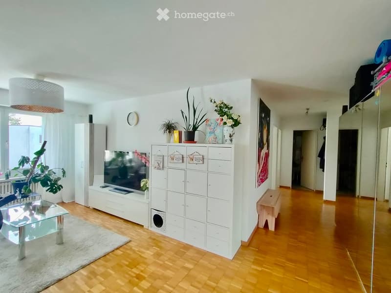 Bright living room with wooden flooring, white walls, a glass coffee table, a TV on a white cabinet, a bench, and several potted plants.