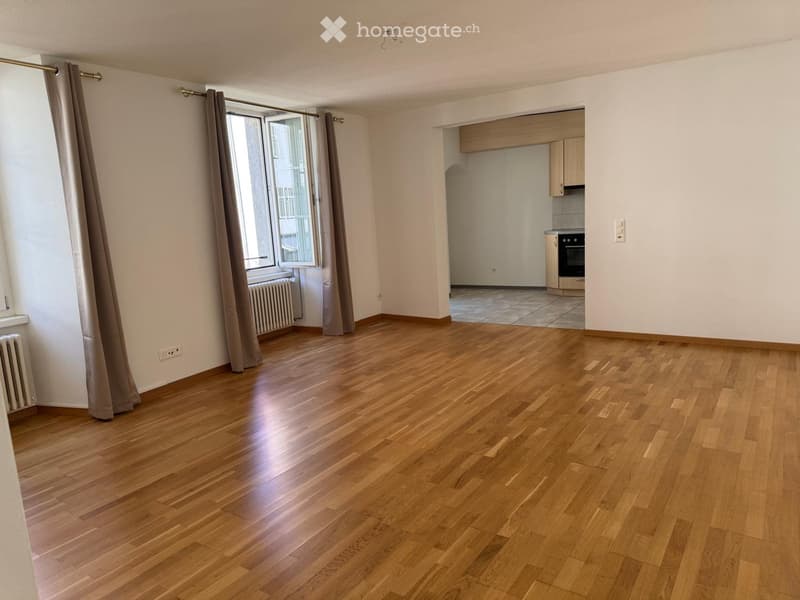 Empty room with wooden flooring, large window with curtains, radiator, and an adjacent kitchen area with cabinets and appliances.