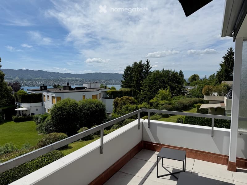 Balcony with a small table, overlooking a landscaped garden and distant lake and mountains.