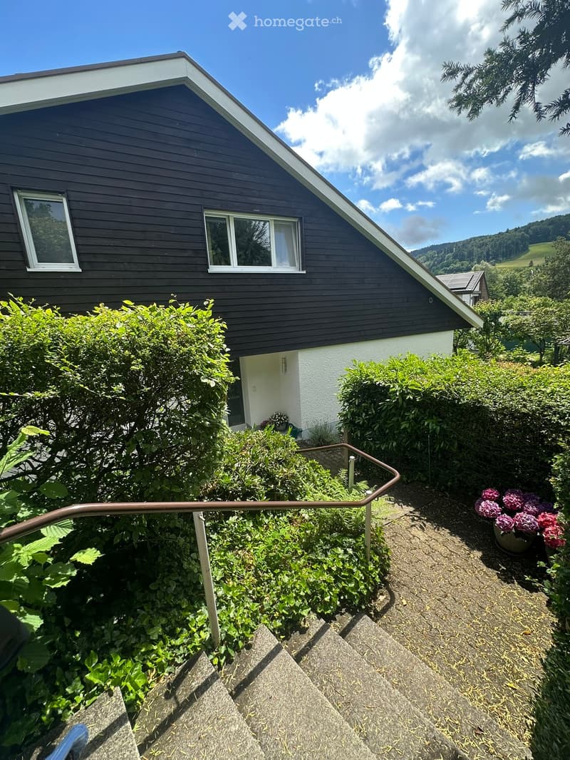Two-story house with dark wooden siding, white trim, and a sloped roof. The house has multiple windows and is surrounded by lush greenery and a staircase leading to the entrance.