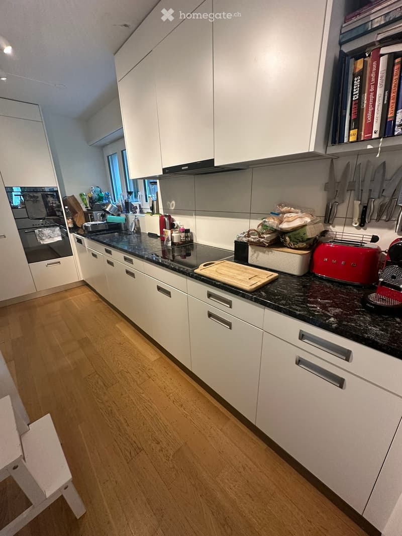 Modern kitchen with white cabinets, dark granite countertops, and wooden flooring. Includes a red toaster, cutting board, knife rack, and various kitchen appliances.