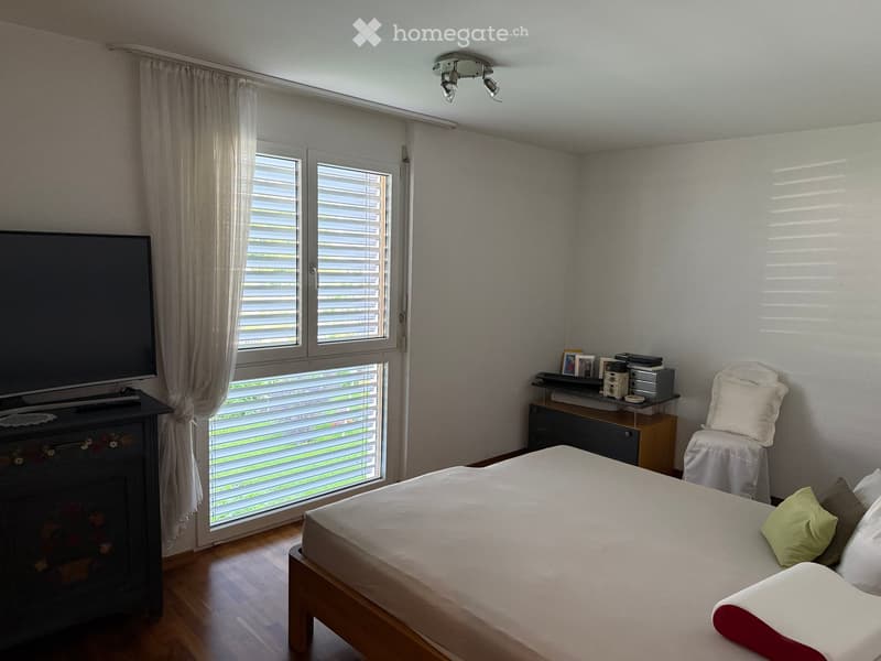 Bright bedroom with wooden flooring, large window with blinds and curtains, bed with pillows and blanket, chair, small table with items, and television on a stand.