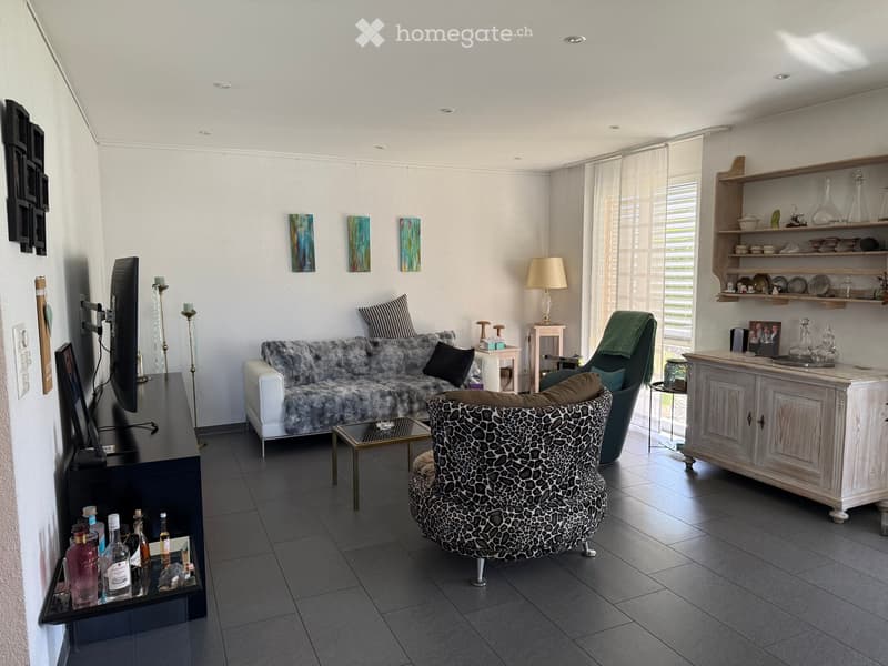 Modern living room with a white sofa, leopard-print chair, coffee table, table lamp, shelves, and a television. The room features large windows with blinds and a dark tiled floor.