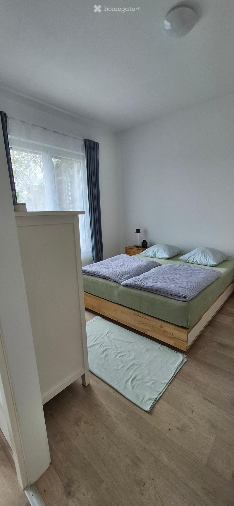 Cozy bedroom with a double bed, nightstand, lamp, and a large window with curtains. Wooden flooring and a small rug in front of the bed.