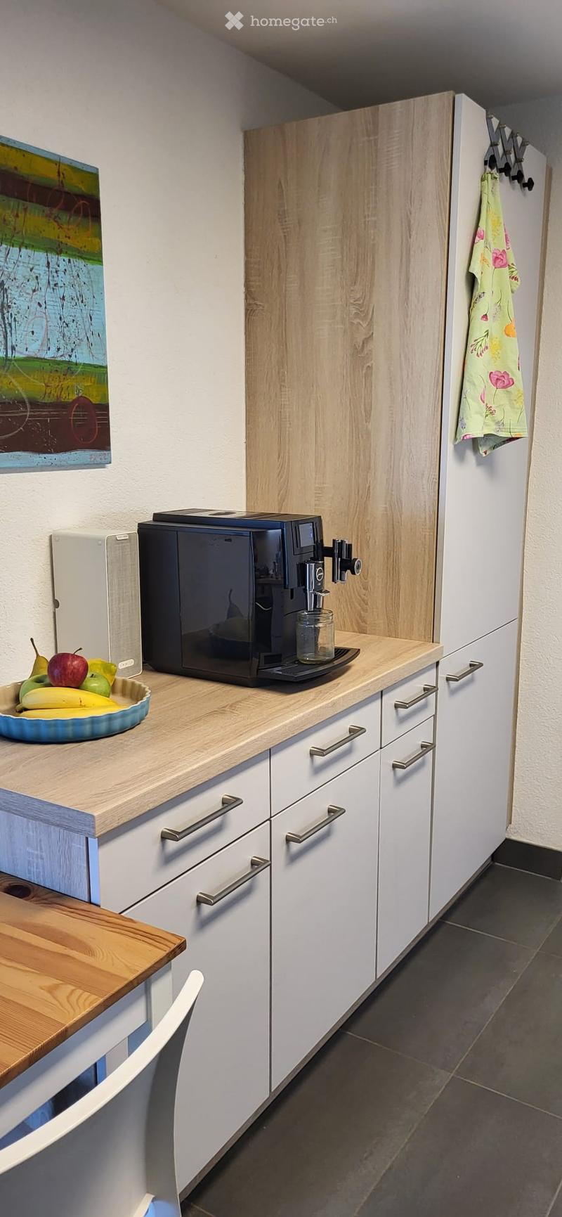 Modern kitchen with white cabinets, wooden countertops, a coffee machine, a fruit bowl, and a small dining area with a table and chair.