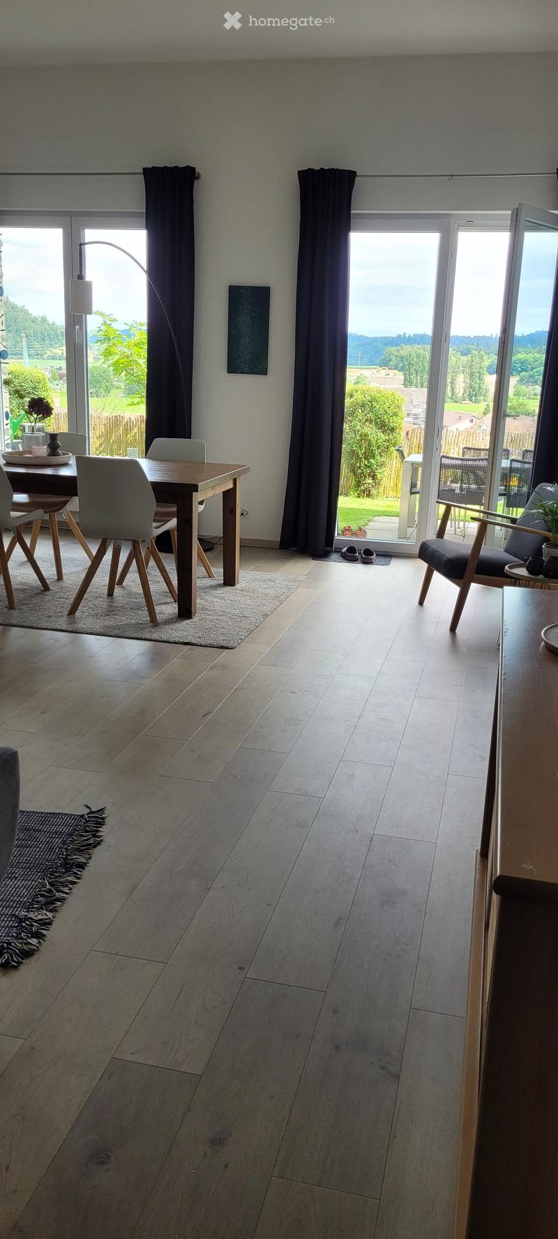 Bright, open living and dining area with wooden flooring, large windows, and a view of the garden. Features a dining table with chairs, a sofa, a table lamp, and a small art piece on the wall.