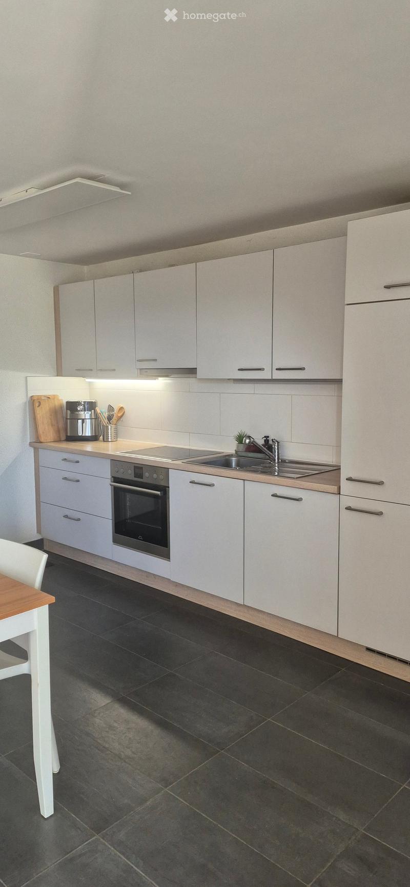 Modern kitchen with white cabinets, dark tiled floor, stainless steel sink, oven, and countertop items including a coffee maker and cutting board.
