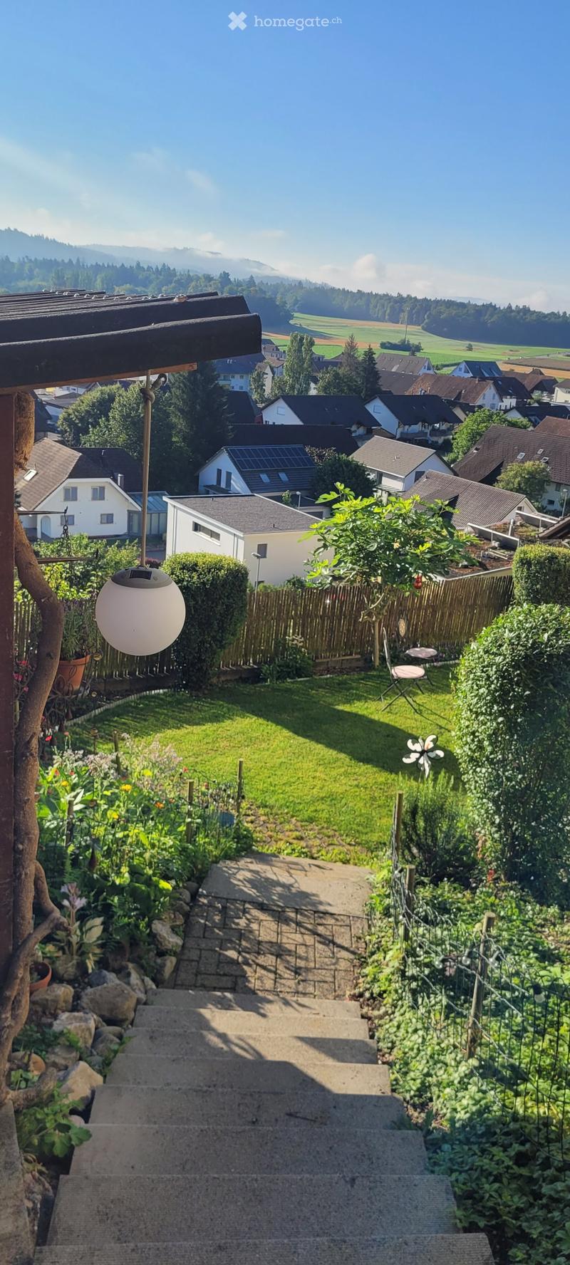 A well-maintained garden with a staircase leading up, surrounded by greenery, flowers, and a small seating area with a table and chairs. The background shows a residential neighborhood with houses and a scenic mountain view.