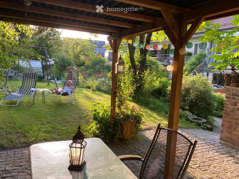 Garden with a covered patio area, outdoor table and chairs, lantern, swing, and lush greenery.