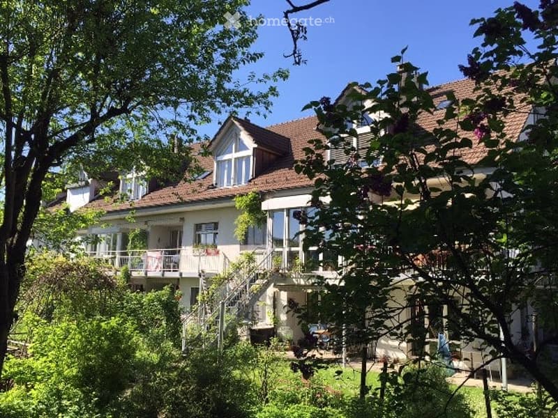 A two-story house with a red-tiled roof, large windows, and a balcony. The house is surrounded by lush greenery and trees.