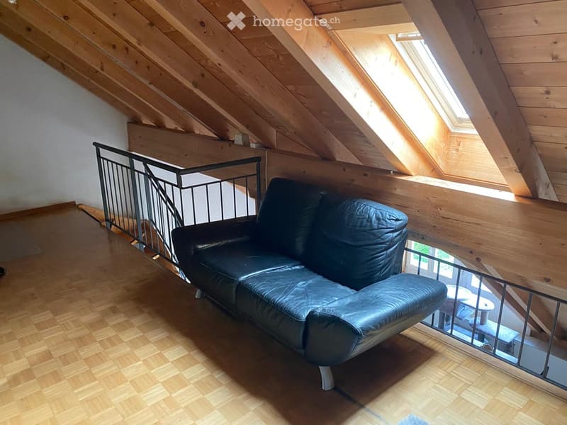 Loft-style room with wooden slanted ceilings, skylight windows, and a black leather sofa. Wooden floor and metal railing visible.