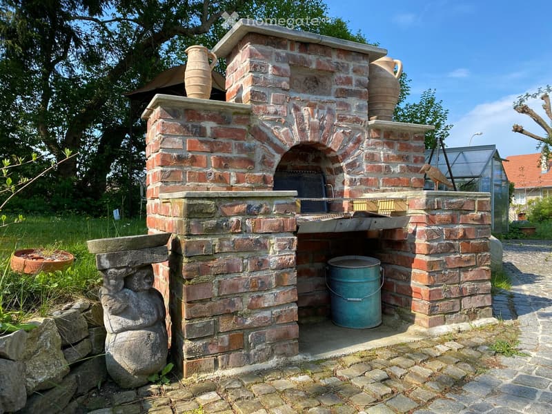 Brick outdoor oven with ceramic vases on top, surrounded by greenery and a stone pathway.