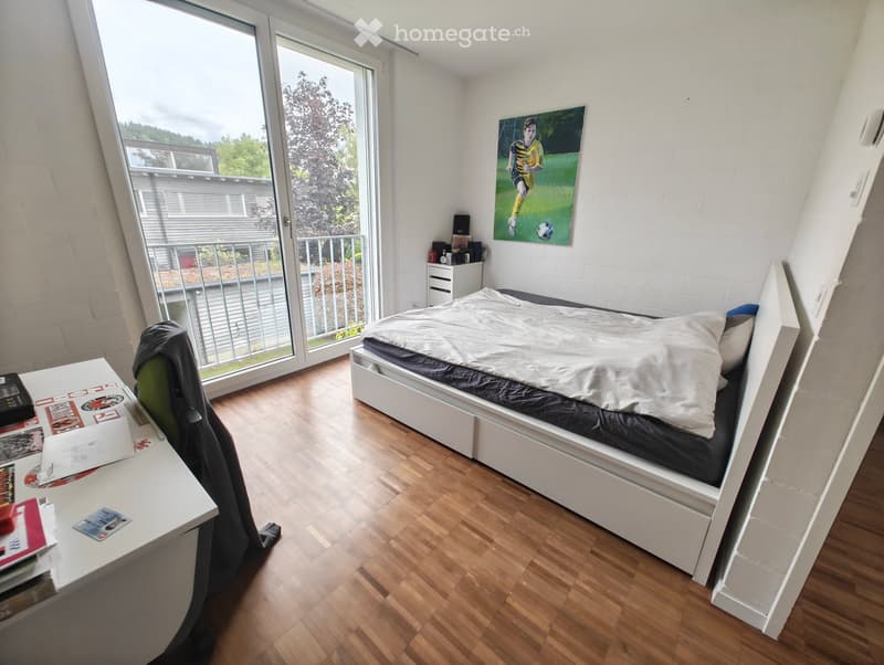 A modern bedroom with a white bed, a desk with a chair, a nightstand, and a large window with a view of the balcony and greenery outside.