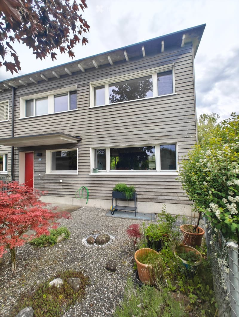 Two-story house with grey wooden siding, multiple windows, and a red front door. The front yard features gravel, potted plants, and a small garden area.