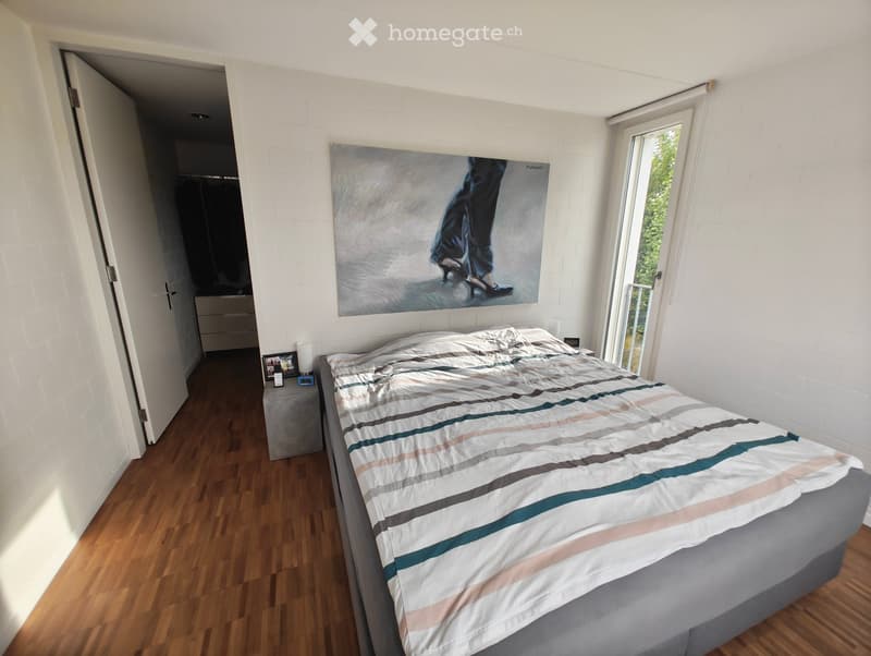 Modern bedroom with a large bed, striped duvet, nightstand with lamp, picture frame, and an open closet area visible in the background.