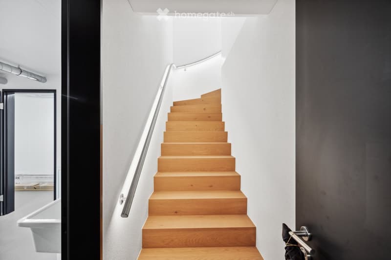 Modern staircase with wooden steps and a sleek metal railing, set against white walls and a dark accent wall, with a glass door visible on the left side.