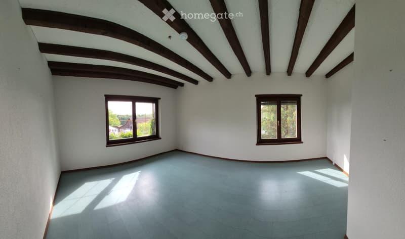 Empty room with white walls, wooden ceiling beams, and two windows with brown frames. The floor is light blue tiles, and the room has a simple, clean design with no furniture or decorations.