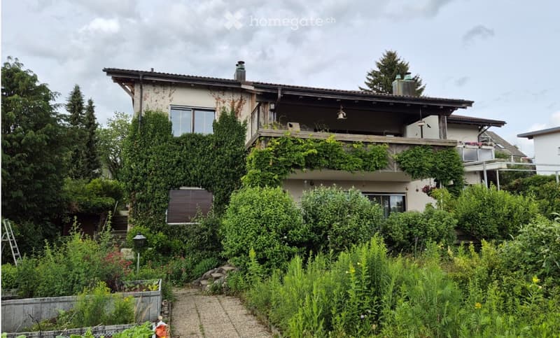 Two-story house with a balcony, surrounded by lush greenery and trees.