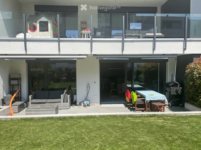 Well-maintained garden with a lawn, patio seating area, outdoor sofa, table, chairs, and a small toy house visible through the glass balcony.