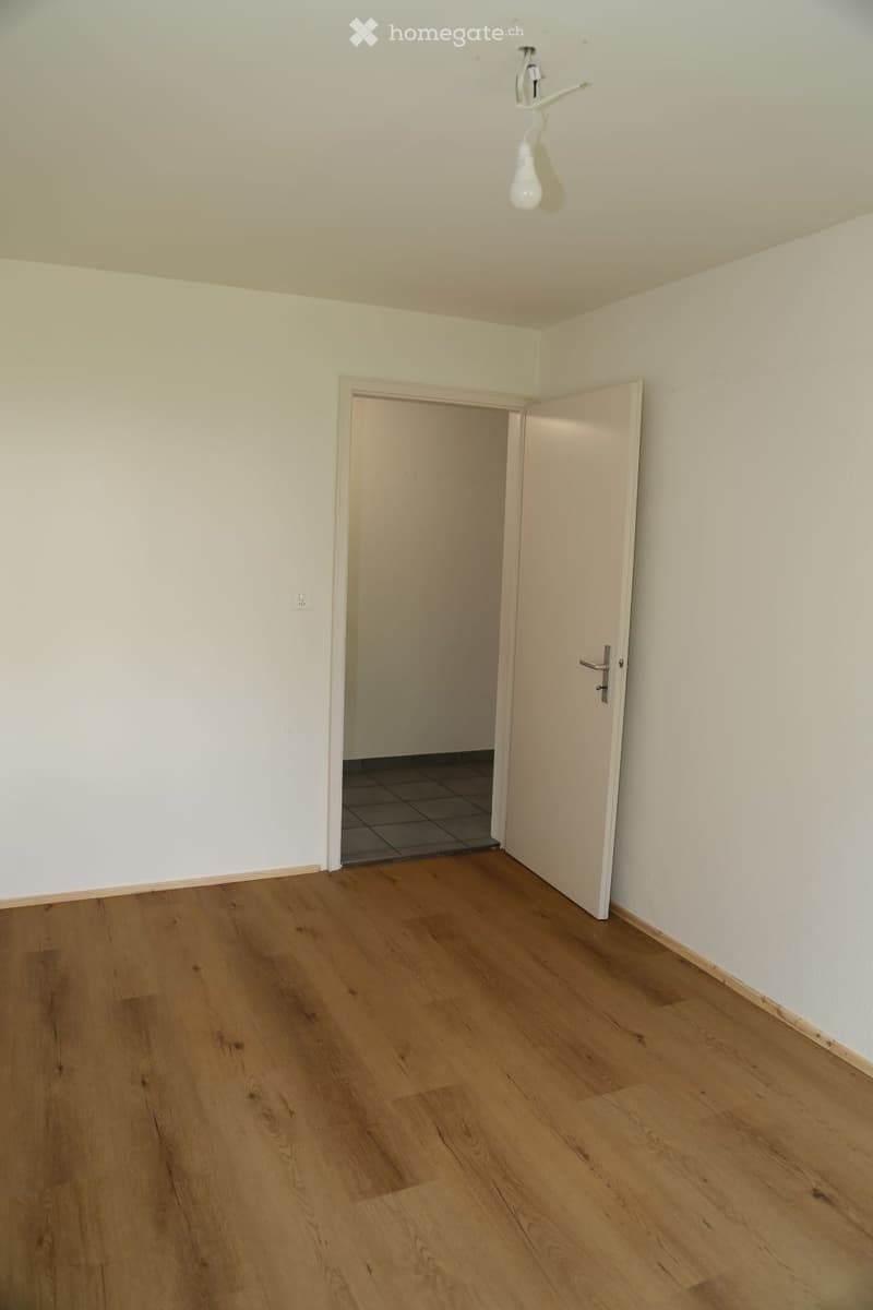 Empty room with light wooden flooring, white walls, and a single open door leading to another room with tiled flooring.