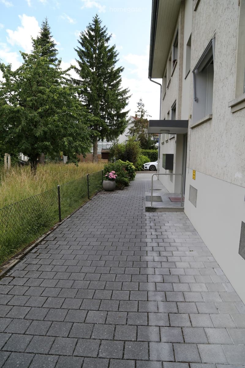 Brick-paved pathway leading to a building entrance, flanked by greenery and a small planter with pink flowers.