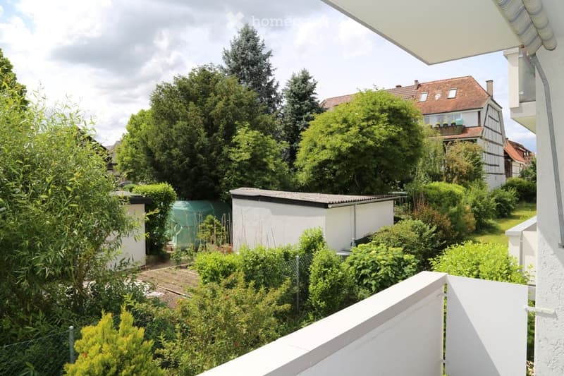 Balcony view of a lush garden with various trees, shrubs, and a small shed. A neighboring house with a balcony is visible in the background.