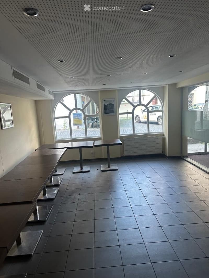 Empty room with tiled floor, several tables and chairs, large arched windows, and a radiator on the wall.