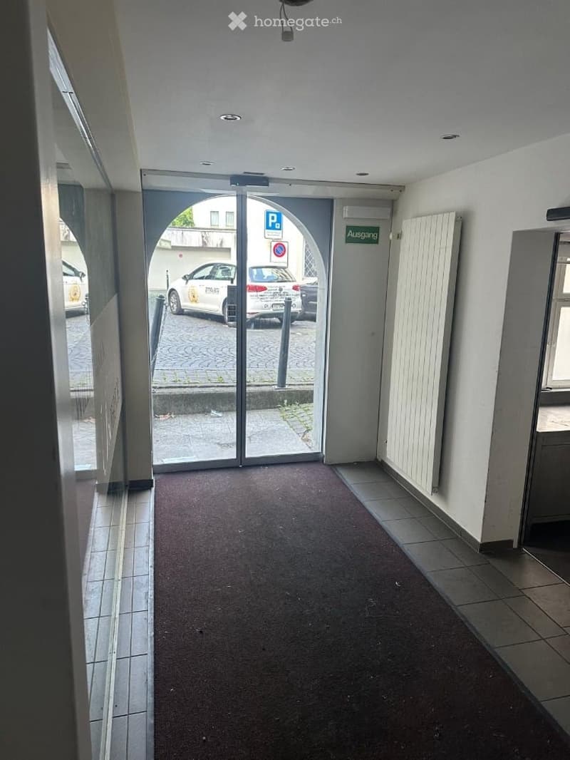Clean, modern hallway with tiled walls, carpeted floor, and a glass automatic door leading to a parking area.