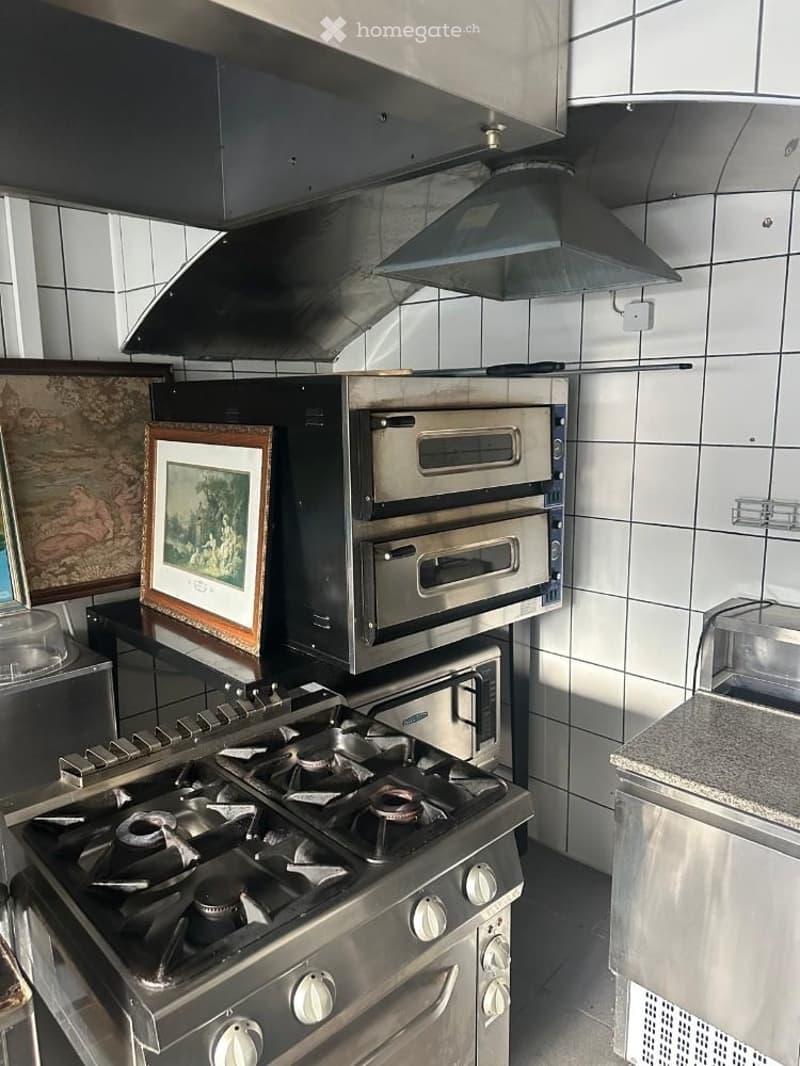 Commercial kitchen with stainless steel appliances, including a gas stove, multiple ovens, and a large range hood. Two framed pictures are placed on the counter.
