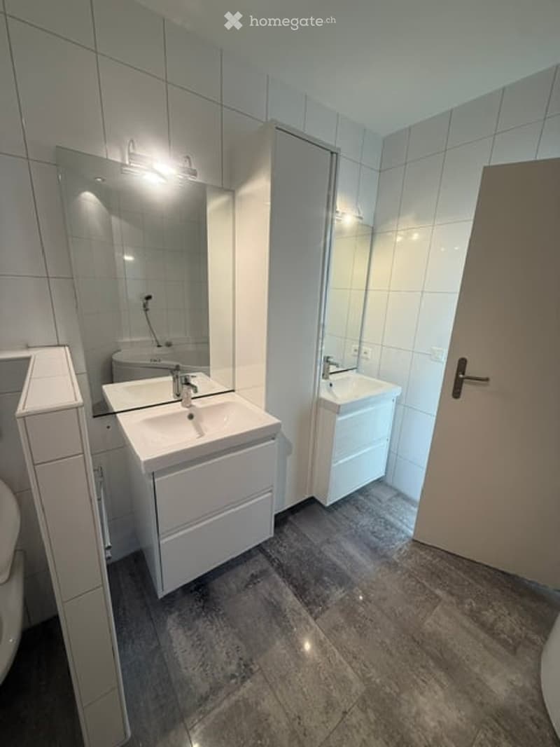 Modern bathroom with white tiled walls, two white sinks with modern faucets, large mirrors, and built-in cabinets.