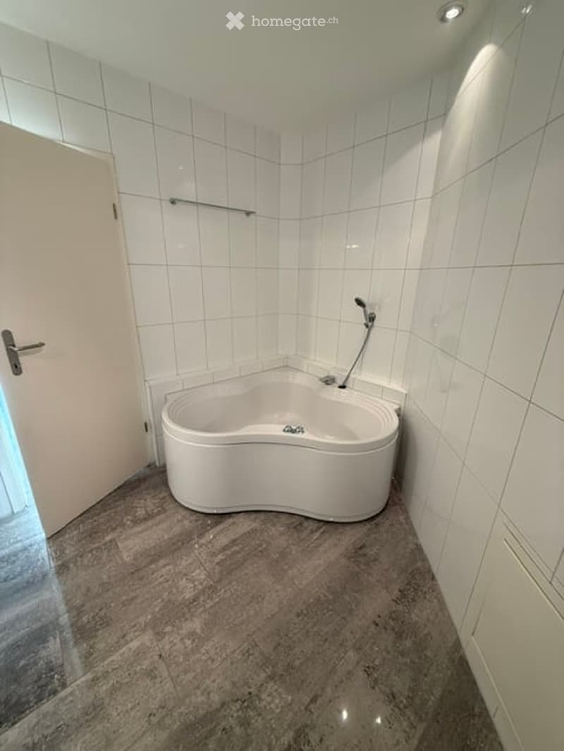 Modern bathroom with a white, curved bathtub, tiled walls, and a marble floor. A door is visible on the left, and a shelf is mounted on the wall above the bathtub.