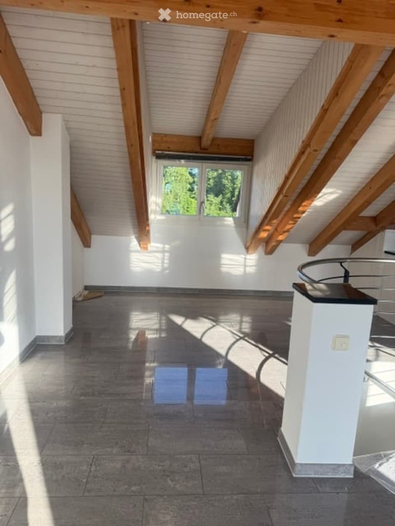 Empty attic room with exposed wooden beams, white walls, and a large window letting in natural light. The floor is covered with dark tiles, and there is a small white column in the corner.