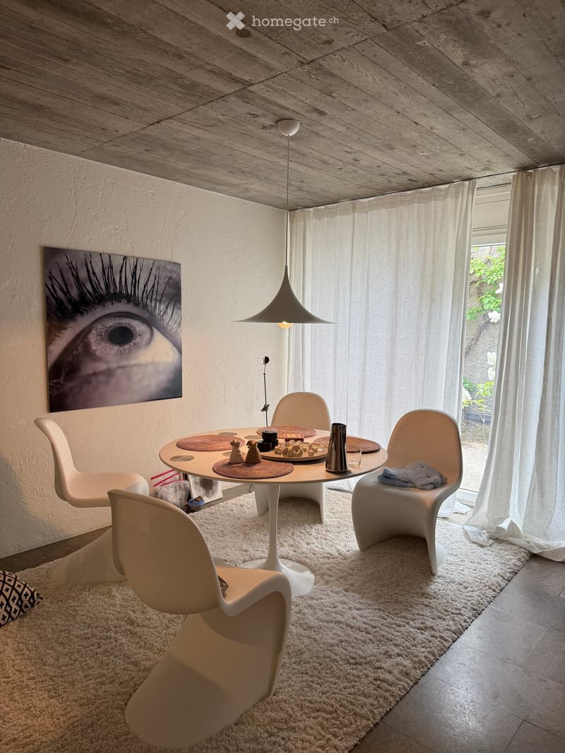 A modern dining area with a round white table, four white chairs, a hanging lamp, and a large eye artwork on the wall. The space features a white shag rug and sheer white curtains.