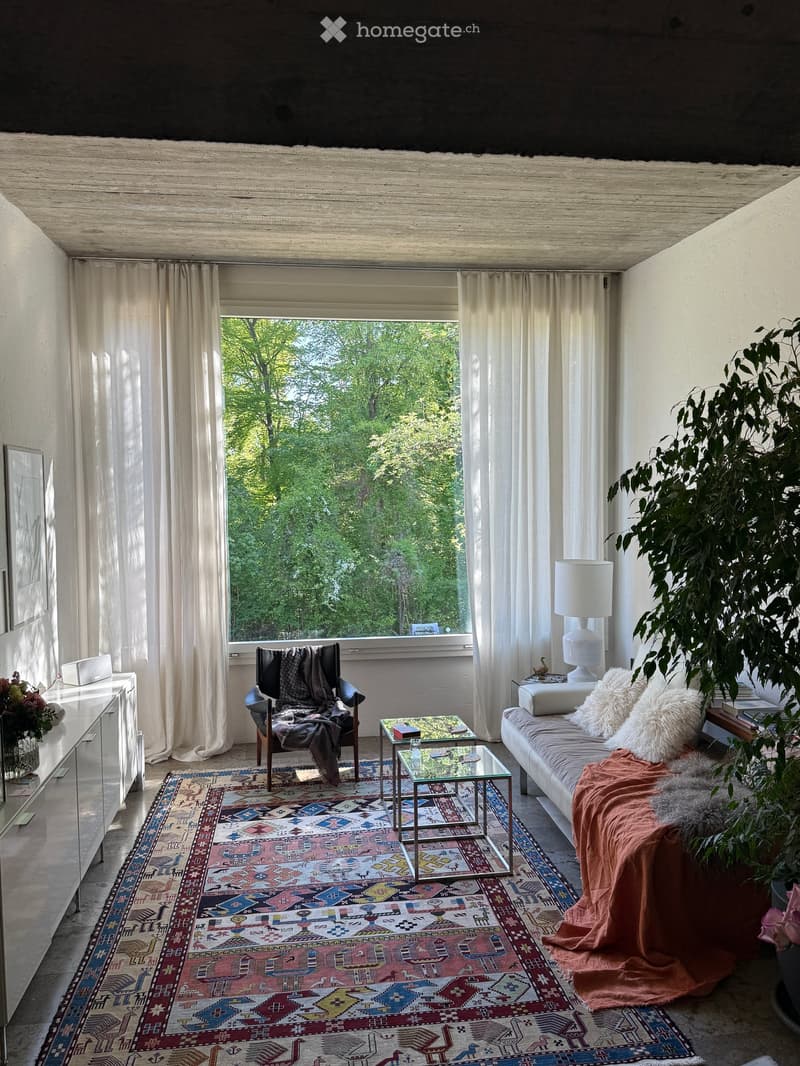 Bright living room with large windows, white curtains, colorful patterned rug, sofa with pillows and blanket, armchair with throw, coffee table, side table with lamp, and decorative items.