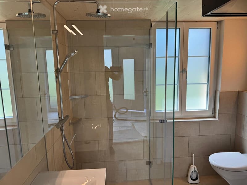 Modern bathroom with a glass-enclosed shower, a bathtub, and a toilet. The shower features multiple heads and a handheld sprayer. The walls are tiled in beige, and the floor is also tiled.