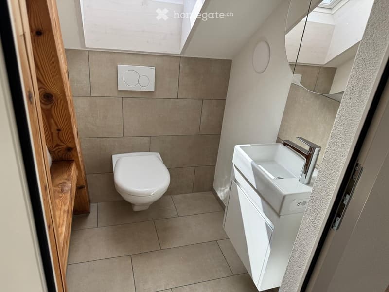 Modern bathroom with a toilet, sink, and cabinet. Features tiled walls and a wooden door frame.