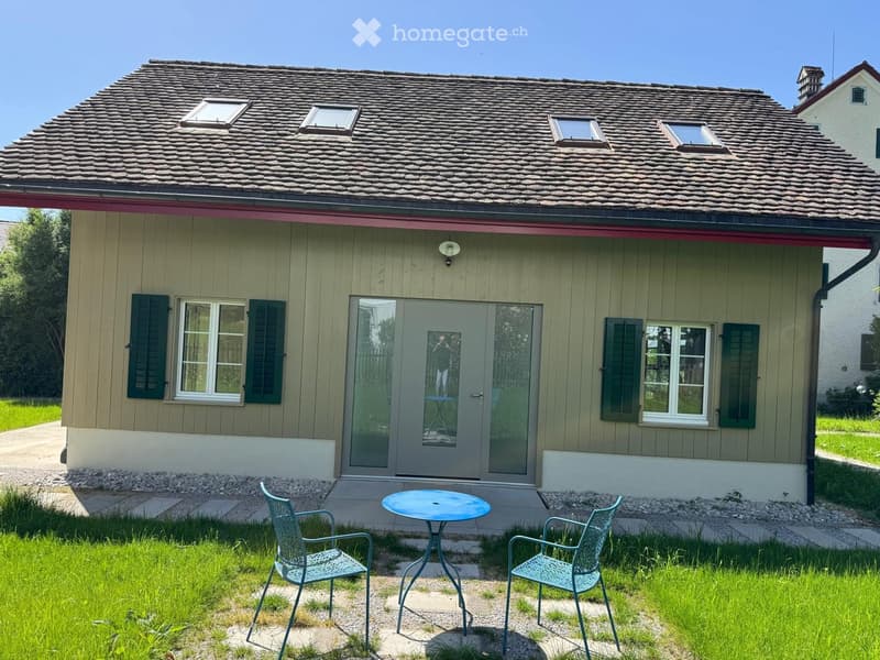 A small, single-story house with a tiled roof and green shutters. A patio area in front features a round table and two matching chairs on a stone pathway.