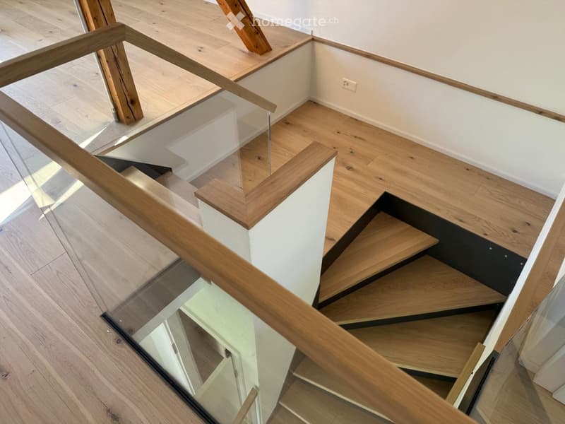 Modern staircase with wooden steps and glass railing, light-colored wooden flooring, white walls, and a visible electrical outlet.