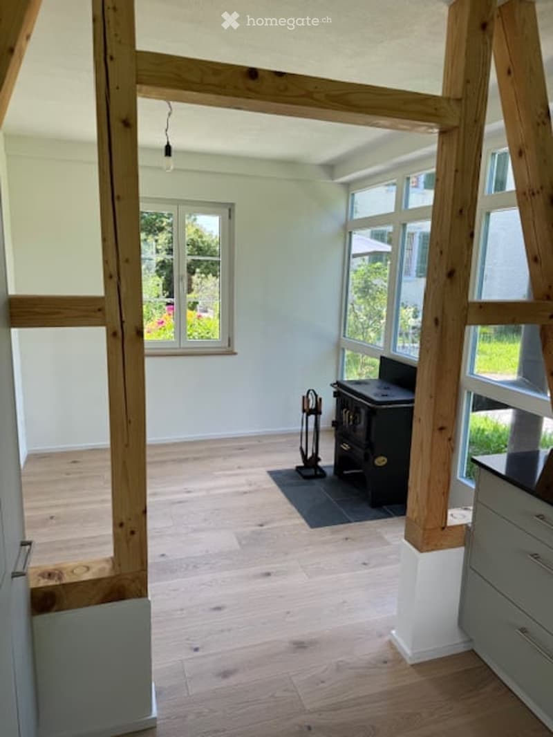 Empty room with wooden flooring, white walls, large windows, and exposed wooden beams. A small black stove and a broom stand are visible, but no furniture.