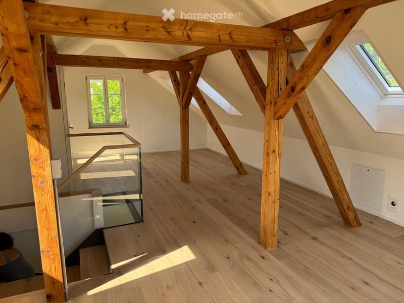 Bright, open attic space with exposed wooden beams, wooden flooring, and skylight windows. Features a glass staircase leading to a lower level.
