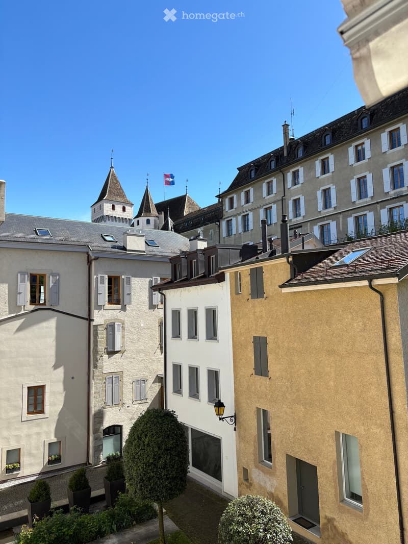 A picturesque European town scene with multi-story buildings, traditional architecture, and a clear blue sky. The buildings feature white and beige facades, shuttered windows, and ornate rooftops. A small garden area with trimmed bushes and potted plants