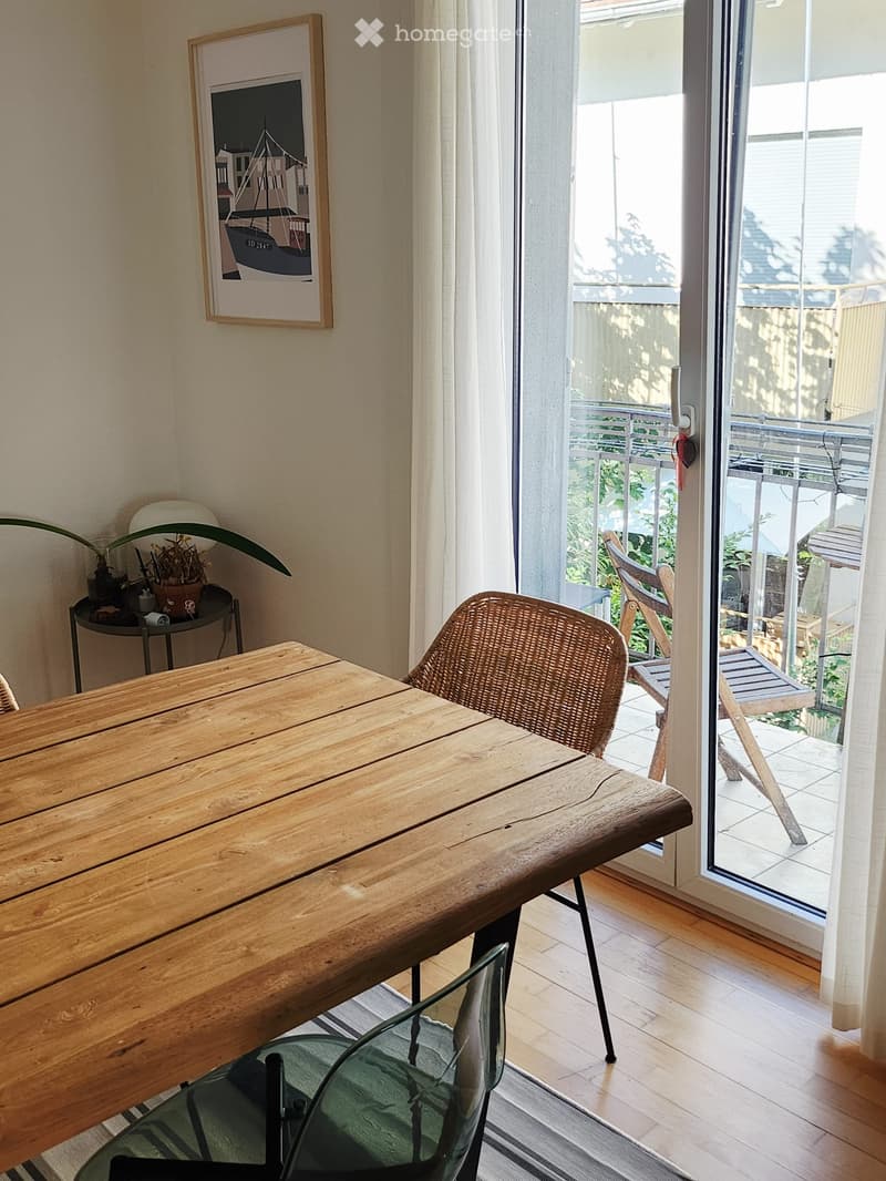 Bright room with wooden floor, large sliding glass door leading to a balcony, wooden table, wicker chair, glass chair, small plant, and picture frame on wall.