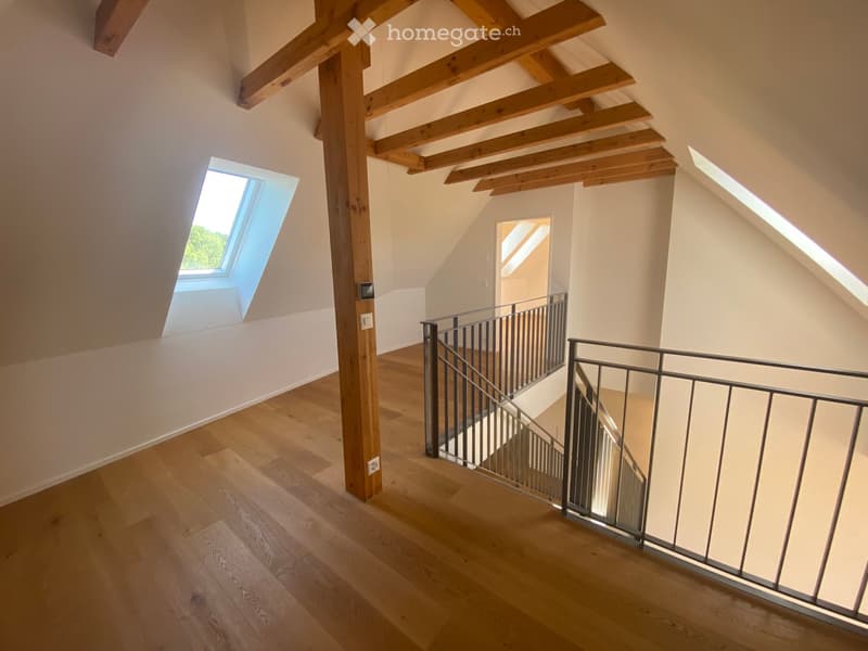 Bright, open hallway with wooden flooring, exposed wooden beams, and a modern metal staircase leading upwards. Skylight windows provide natural light.