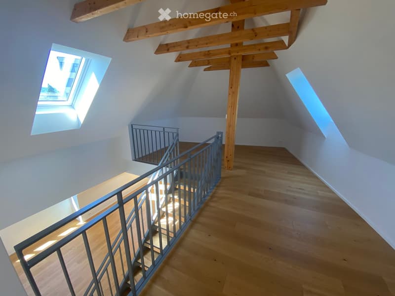Bright, airy hallway with wooden flooring, exposed wooden beams, and a modern metal railing leading to a staircase. Skylights provide natural light.