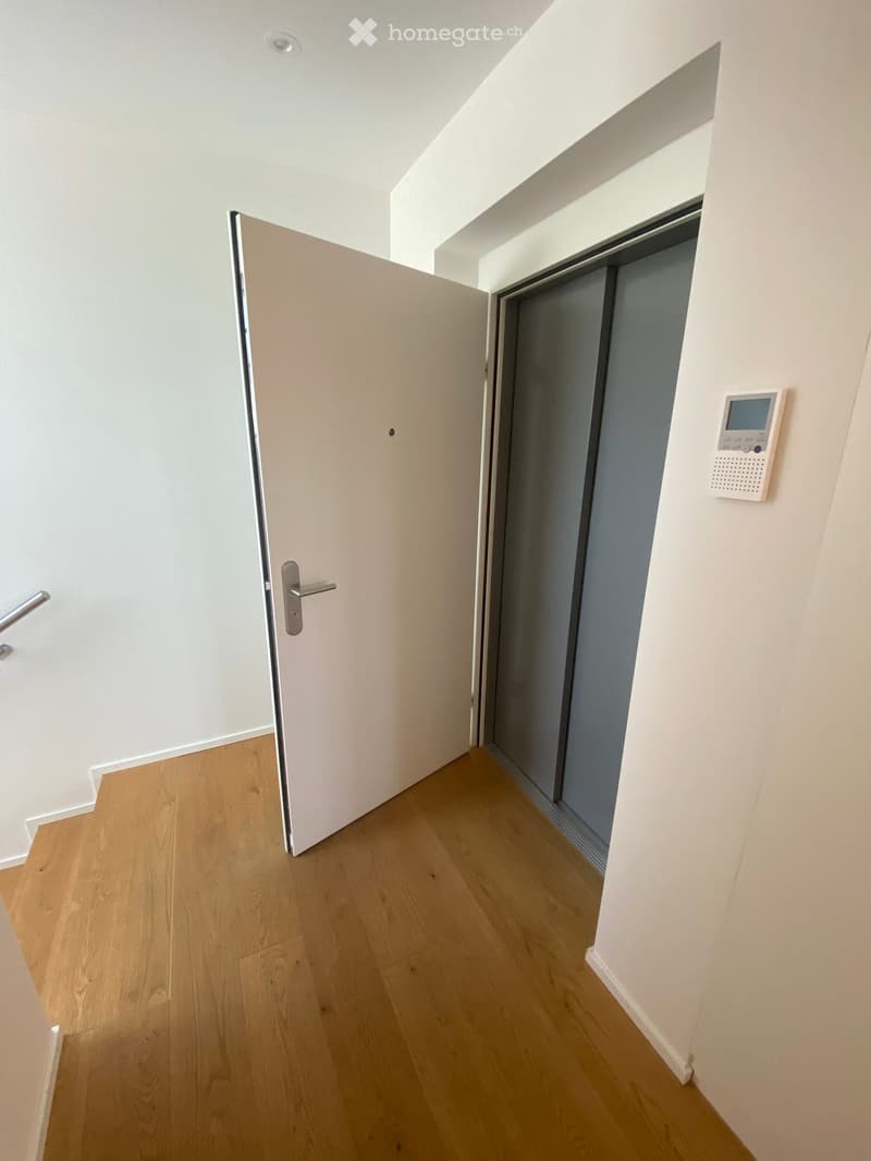 Empty hallway with light wooden flooring, white walls, and a partially open door leading to another room. A control panel is mounted on the wall near the elevator door.